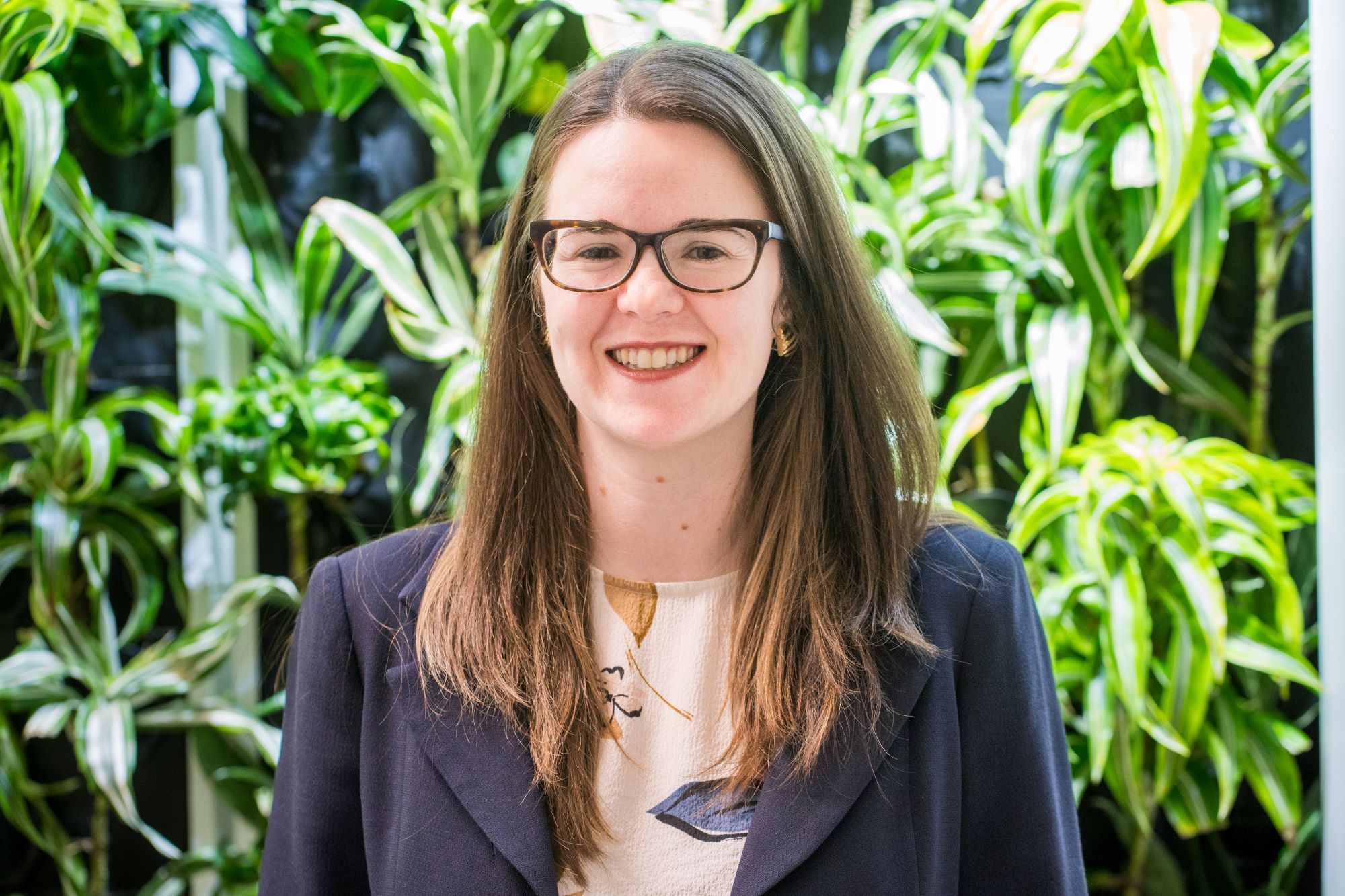 A photo of Catherine Thorn in front of a green wall. She is wearing glasses and smiling.