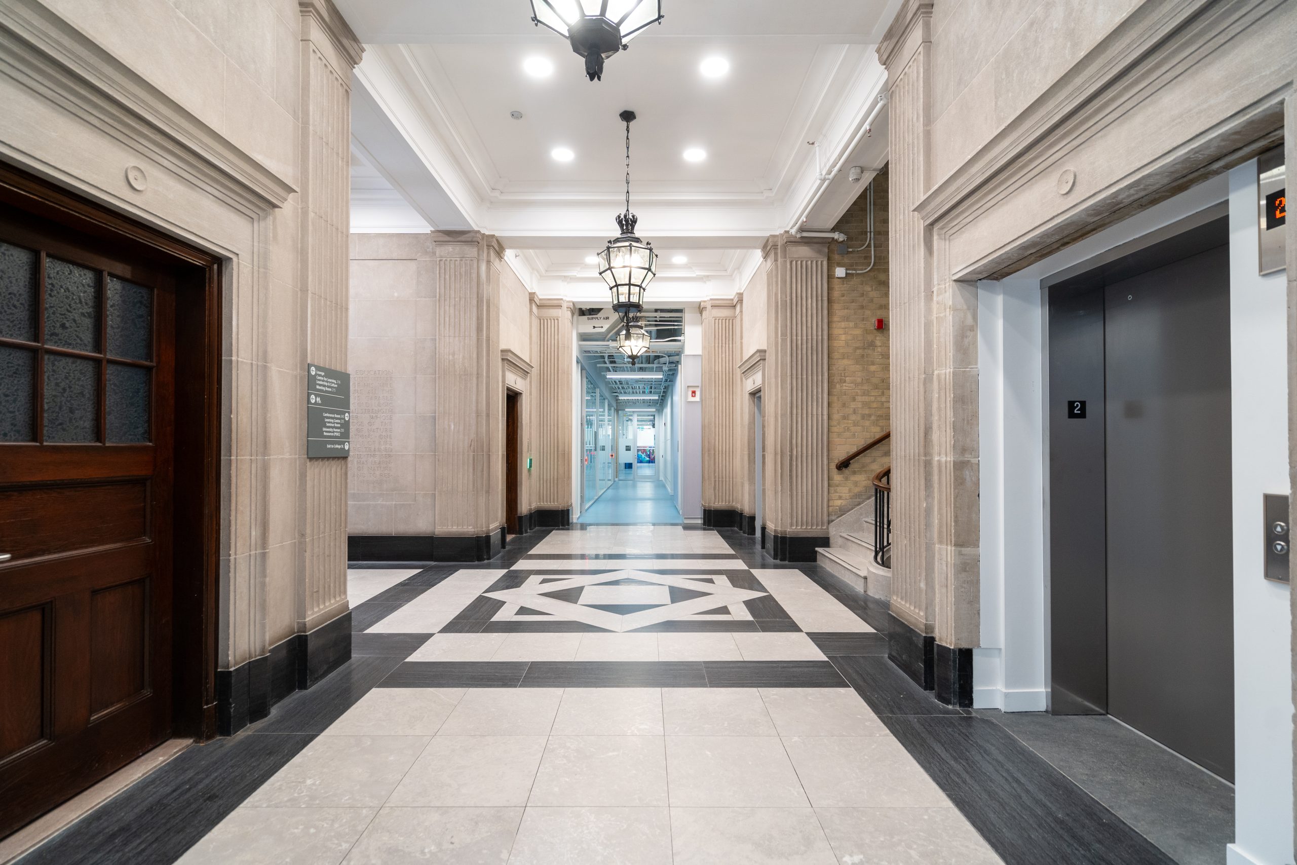 A photo of a hallway in the Fitzgerald Building. It is bright and clean, with tile flooring in a mosaic pattern, and features crown molding, historical looking hanging lights and ionic pillar design, among other such ornate details.